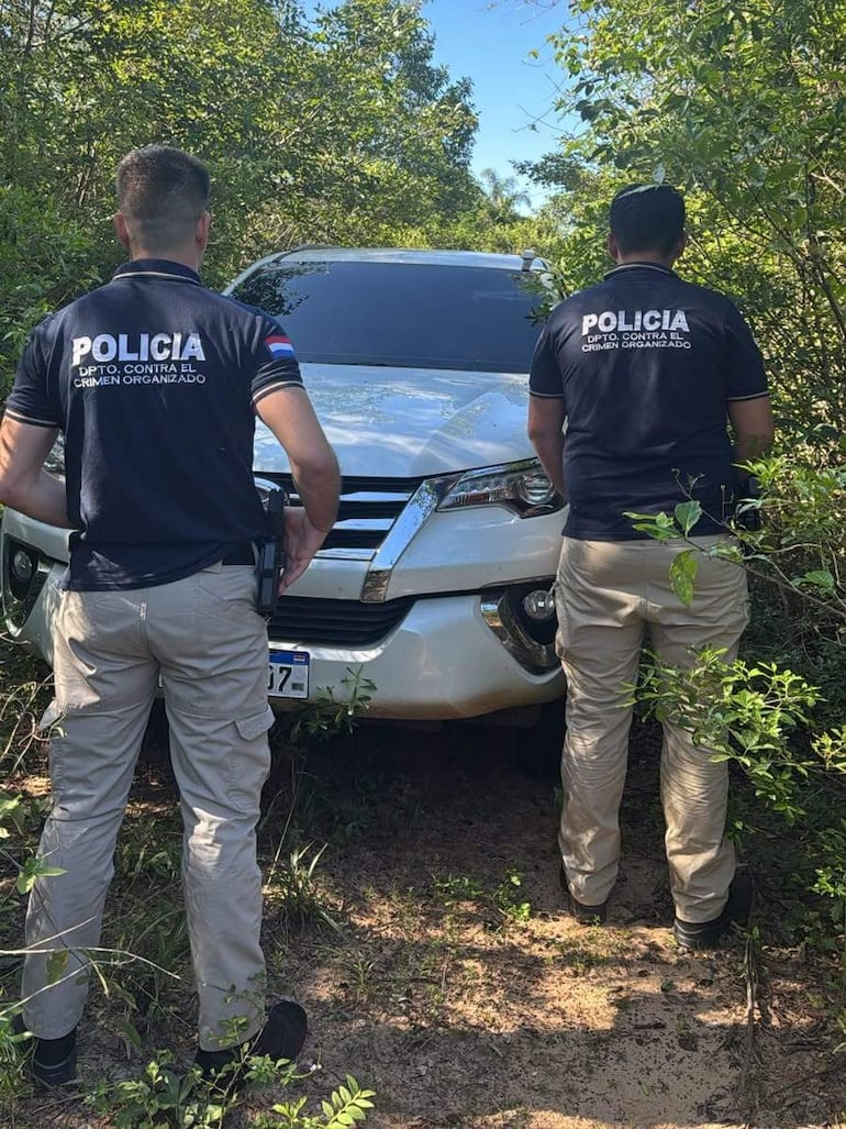 Dos agentes de policía de espaldas, vestidos con camisetas oscuras, observan una SUV blanca cubierta de maleza en un entorno natural.