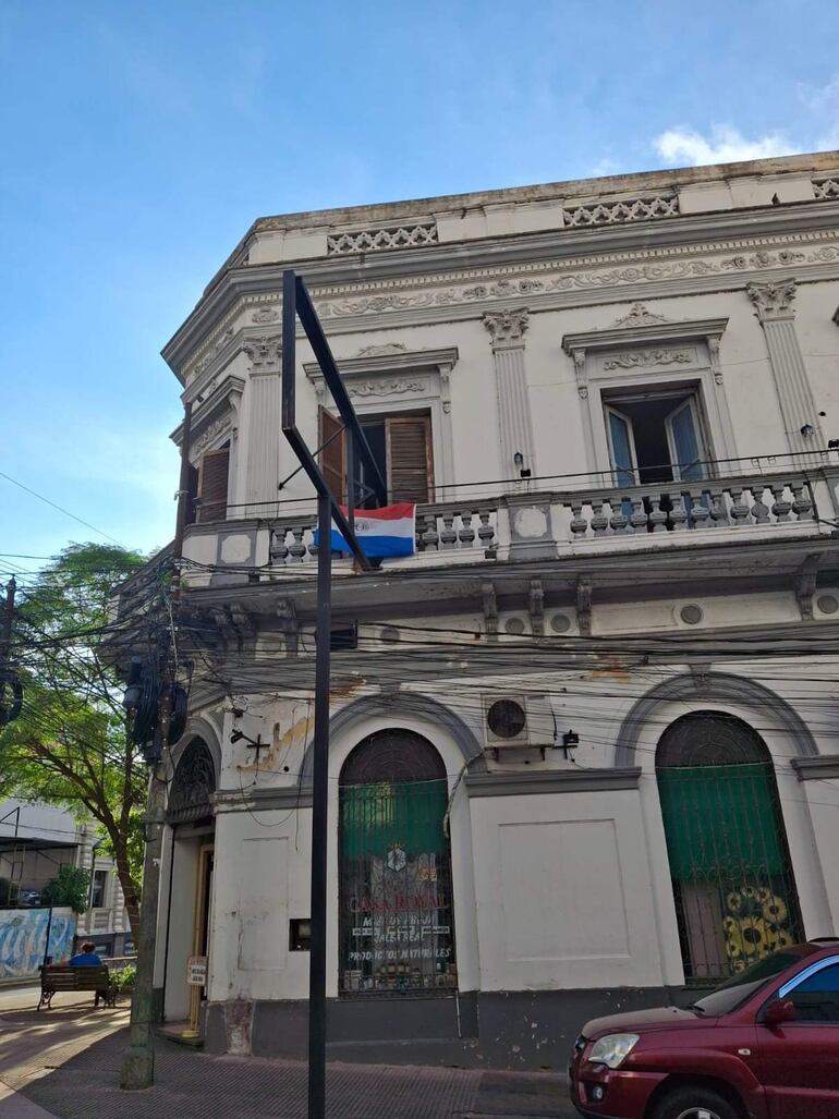 Edificio colonial en Asunción con asta de bandera paraguaya, sin personas visibles y vehículo rojo estacionado.
