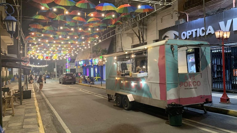 Camión de comida 'Avenida Porco' estacionado frente a la Universidad Americana, rodeado de luces decorativas y personas socializando.