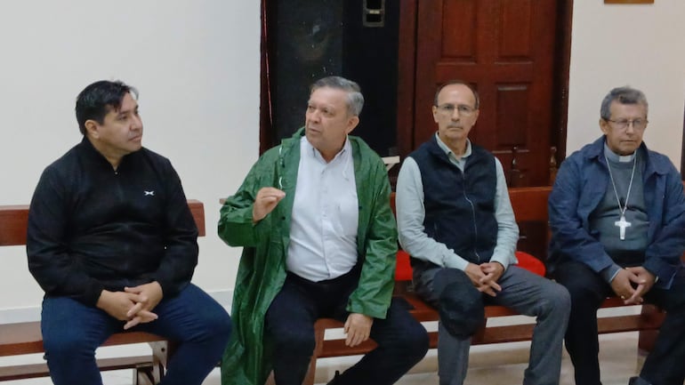 Cuatro hombres en una reunión interior. Uno habla con chaqueta verde, mientras los demás escuchan atentos con diferentes atuendos.