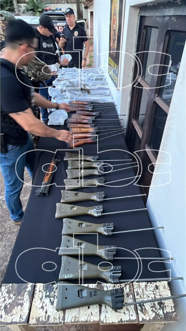 Cuatro agentes con uniformes oscuros junto a una mesa con rifles y un emblema institucional en el fondo.