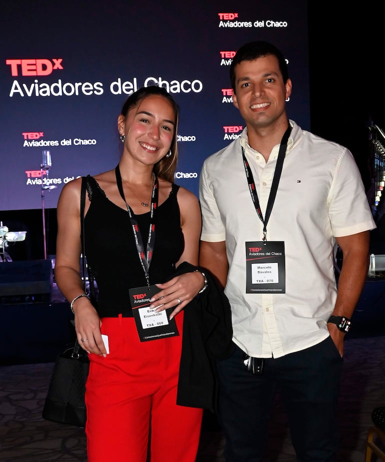 Mujer con blusa negra y pantalones rojos sonríe mientras sostiene una bolsa, junto a un hombre con camisa clara en evento TEDx.
