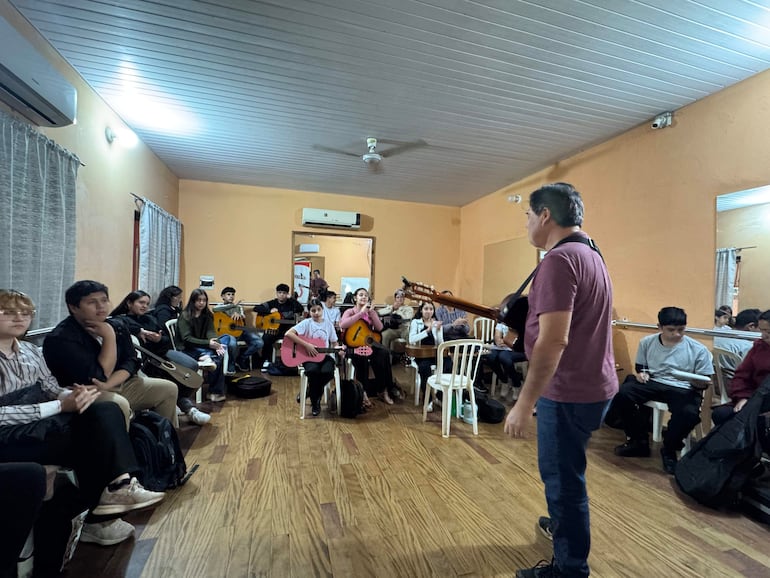 Víctor Riveros, de pie con guitarra, instruye a un grupo de jóvenes sentados con sus instrumentos en un salón de madera.