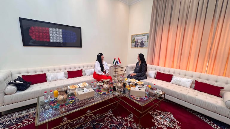 Dos mujeres conversan sentadas en un sofá; una viste blusa blanca y falda roja, la otra en atuendo gris, en un entorno decorado.