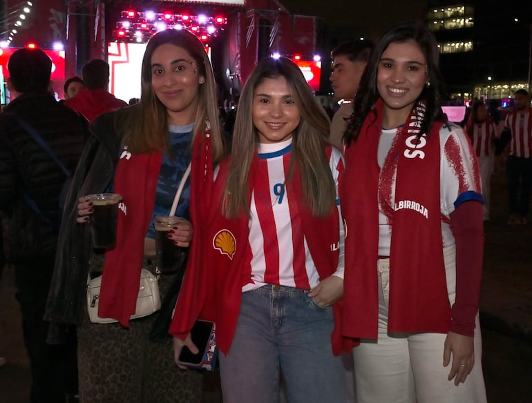 Tres mujeres sonrientes en camiseta y bufanda, posan felices con vasos en un ambiente festivo y de celebración.