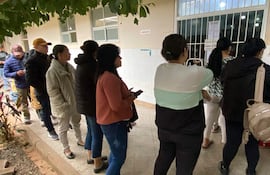 Diez personas con abrigos esperan en fila frente a una ventanilla, sosteniendo teléfonos móviles en un ambiente de farmacia.