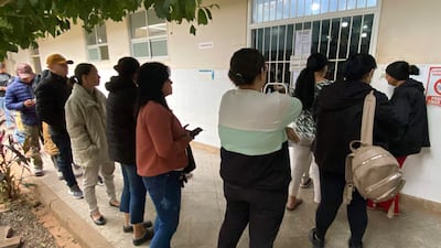 Diez personas con abrigos esperan en fila frente a una ventanilla, sosteniendo teléfonos móviles en un ambiente de farmacia.