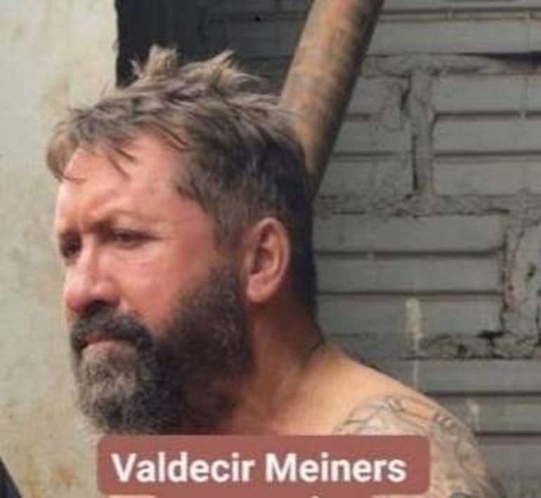 Valdecir Meiners, con barba y cabello corto, tiene expresión seria frente a una pared de ladrillos en un entorno urbano.