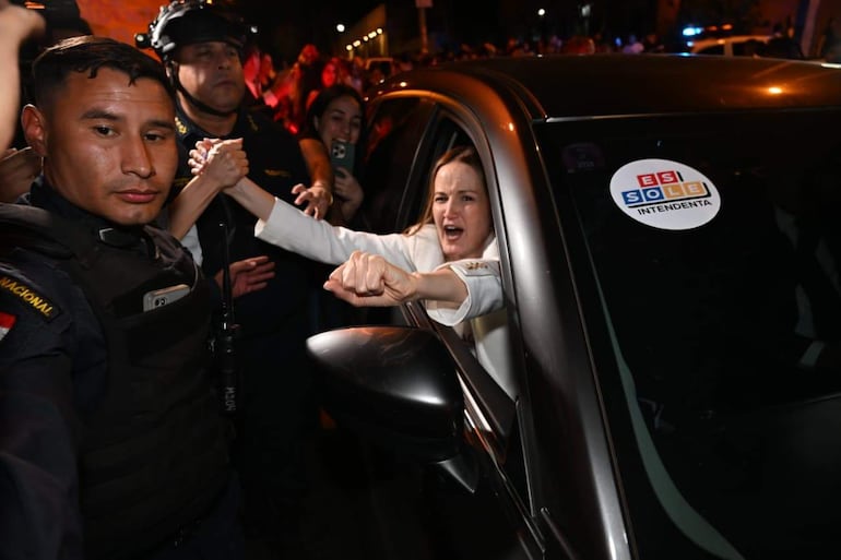 Soledad Núñez en chaqueta blanca asomada por la ventana de un auto, sonriendo mientras seguidores intentan acercarse.