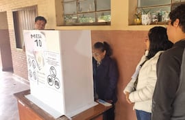 Mujer mayor con gafas vota concentrada, asistida por otra mujer en chaqueta blanca, mientras un hombre supervisa.