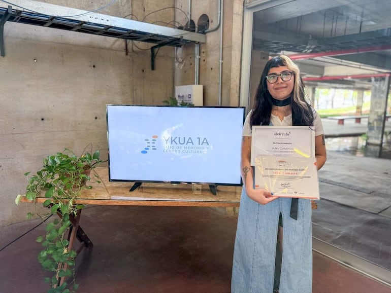 Iara Campos, con blusa blanca y falda celeste, sonríe al sostener un reconocimiento en un ambiente moderno con pantallas y plantas.