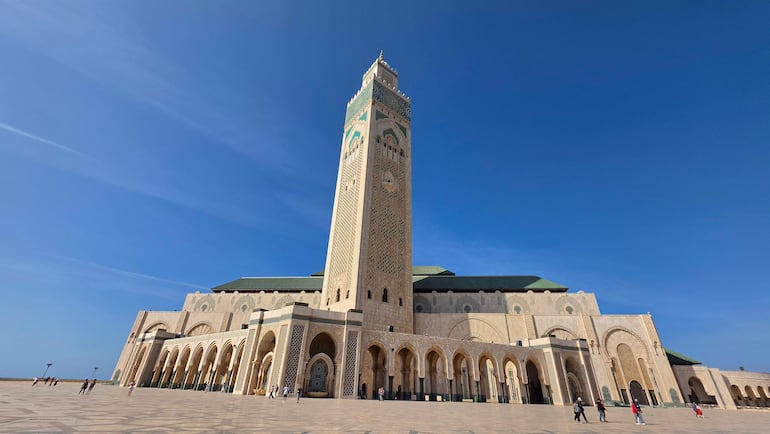Gran Mezquita de Hassan II con torre decorada, rodeada de visitantes dispersos en un ambiente claro y soleado.