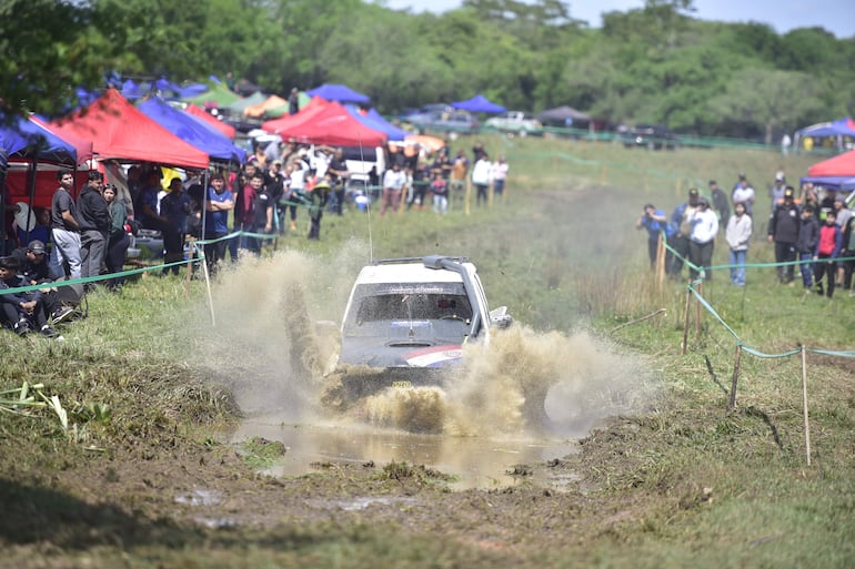 Un coche 4x4 en acción, levantando barro y agua, con un grupo de espectadores entusiastas observando alrededor.