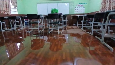 Aula vacía con suelos de madera dañados, escritorios y sillas alineados. Carteles 'PERSONAL' y 'SOCIAL' visibles en la pared.