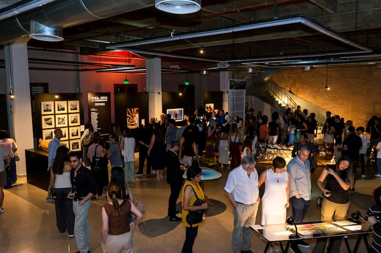 Asistentes conversan y observan fotografías en la inauguración, en un ambiente social con decoración moderna y luz tenue.