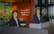 Dos mujeres sonríen en el mostrador de recepción con chaquetas oscuras y blusas blancas, rodeadas de vegetación y un letrero de bienvenida.