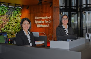 Dos mujeres sonríen en el mostrador de recepción con chaquetas oscuras y blusas blancas, rodeadas de vegetación y un letrero de bienvenida.