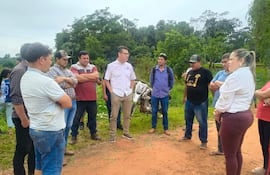 Siete personas, una mujer al frente y hombres escuchando, en un entorno rural con vegetación y ropa informal en tonos variados.