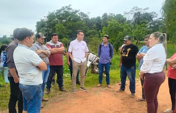 Siete personas, una mujer al frente y hombres escuchando, en un entorno rural con vegetación y ropa informal en tonos variados.