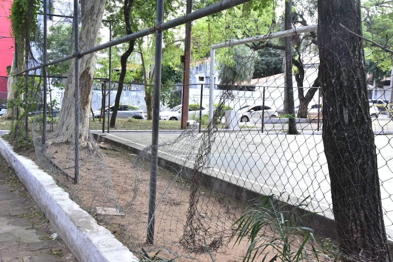 Plaza en mal estado con malla metálica dañada y camino de tierra sin uso. Espacio deportivo vacío y tranquilo.