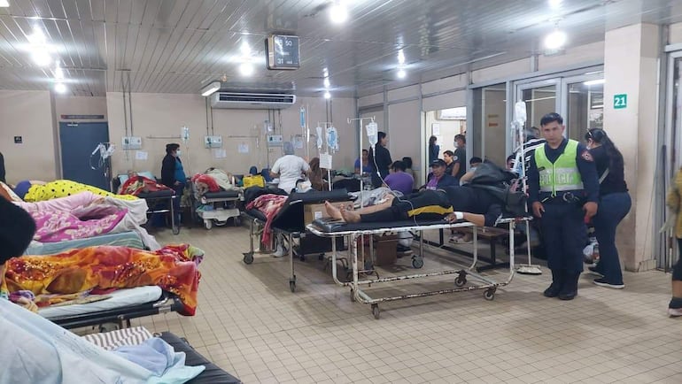 Hombre en camilla con suero en hospital abarrotado, mientras un policía observa atento y personal de salud asiste a otros pacientes.