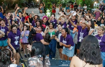 Grupo de mujeres sonrientes con camisetas moradas, disfrutando en evento comunitario al aire libre con vegetación alrededor.