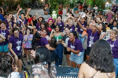 Grupo de mujeres sonrientes con camisetas moradas, disfrutando en evento comunitario al aire libre con vegetación alrededor.
