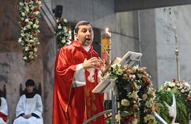 Sacerdote de vestiduras rojas gesticulando en la misa, rodeado de asistentes en vestimenta blanca y decoraciones florales.