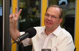 Hombre con camisa blanca y gafas habla sosteniendo un micrófono, fondo amarillo y vaso con agua en la mesa.