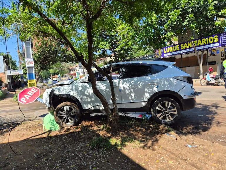 Accidente de tráfico con SUV blanco montado en bordillo junto a árbol y cartel de 'PARE' visible en entorno urbano.