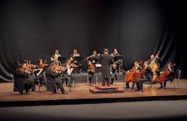 Orquesta de 14 músicos en trajes oscuros, sentados y concentrados, dirigiéndose al director de espaldas en un escenario iluminado.