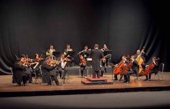 Orquesta de 14 músicos en trajes oscuros, sentados y concentrados, dirigiéndose al director de espaldas en un escenario iluminado.