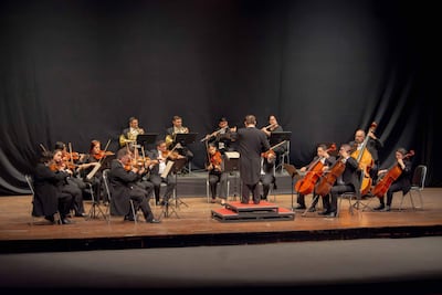 Orquesta de 14 músicos en trajes oscuros, sentados y concentrados, dirigiéndose al director de espaldas en un escenario iluminado.