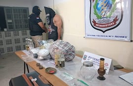 Oficial de policía con chaleco y gorra junto a detenido sin camiseta, con mesas llenas de frascos y bolsas en oficina policial.