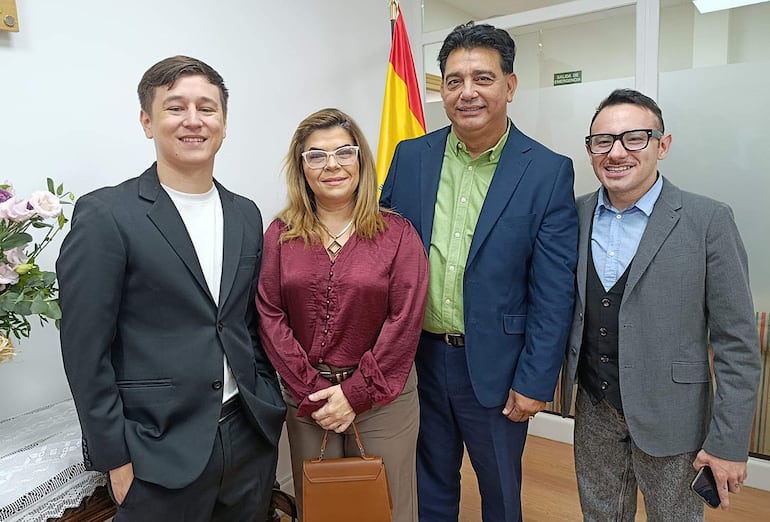 Hombre con blazer negro y camiseta blanca sonríe junto a mujer en blusa burdeos. Ambos interactúan con un hombre con saco azul oscuro y otro con saco gris.