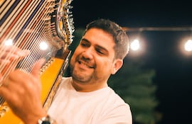 Marcelo Ojeda toca un arpa, sonriente, con camiseta blanca y pulsera, en ambiente festivo con luces tenues.