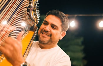 Marcelo Ojeda toca un arpa, sonriente, con camiseta blanca y pulsera, en ambiente festivo con luces tenues.