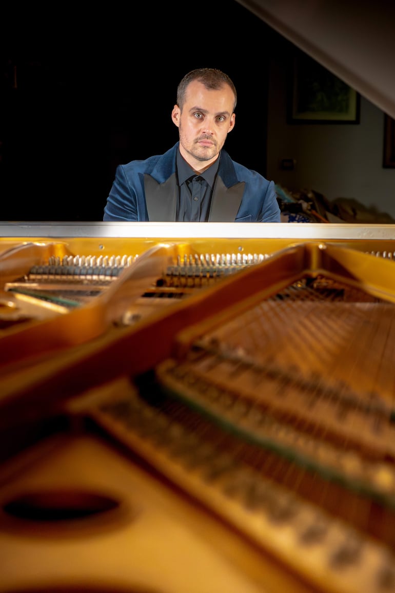 Hombre en traje azul oscuro frente a un piano, concentrado en tocar, con expresión seria y entorno decorativo sutil.
