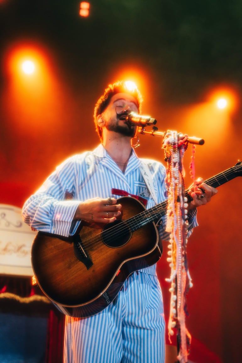 Hombre en pijama de rayas azul y blanco, cantando con guitarra acústica bajo luces cálidas en un escenario.