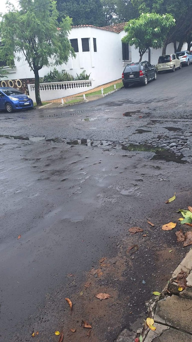 Calle empedrada y húmeda con un vehículo azul estacionado, rodeada de casas clásicas y vegetación otoñal.
