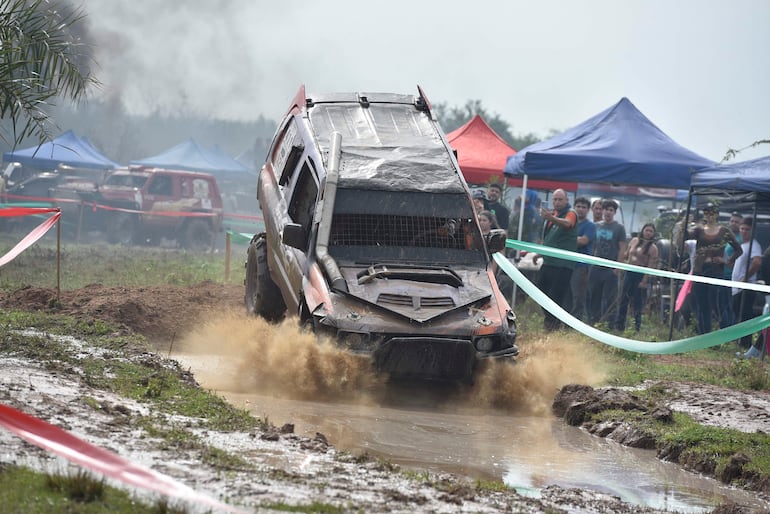 Vehículo todoterreno salpicando barro en competiciones de automovilismo, con espectadores aplaudiendo en carpas de colores.