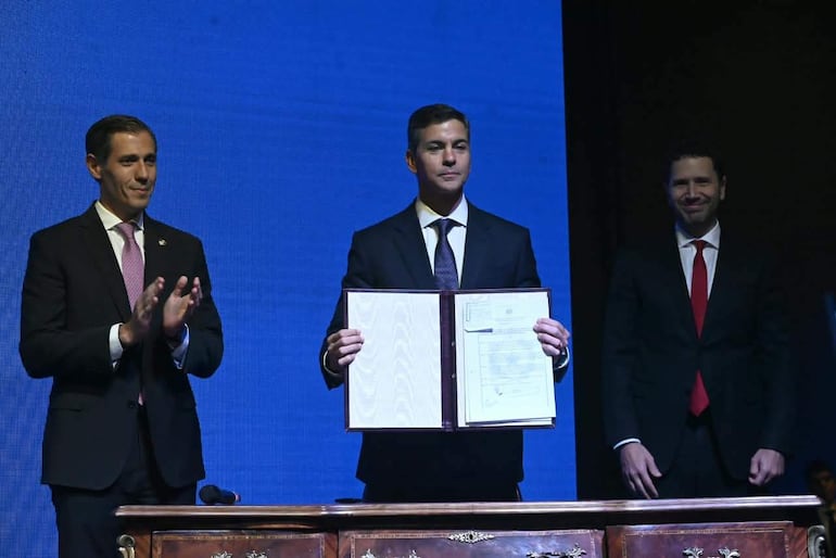 Santiago Peña sonríe mientras presenta un documento, acompañado por otros hombres aplaudiendo en un evento formal con fondo azul.