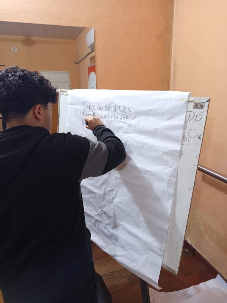 Persona de espaldas con cabello rizado, camiseta negra, escribiendo en caballete en una sala con paredes naranjas.