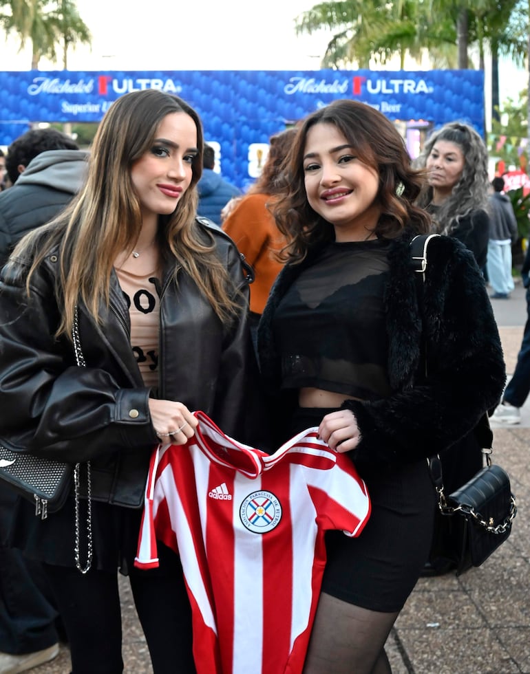 Dos mujeres sonríen al posar juntas, una con chaqueta de cuero y camiseta de fútbol, en un ambiente festivo al aire libre.