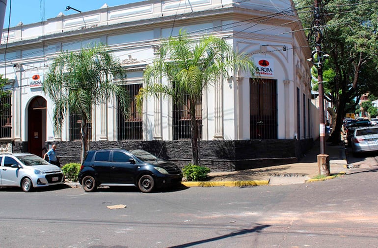 Calle de Aurora con vehículos estacionados y edificio clásico, en un día soleado, sin personas presentes.