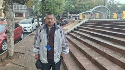 Martín Rojas de pie con chaqueta gris, frente a escalera y vehículos estacionados, en un ambiente urbano.