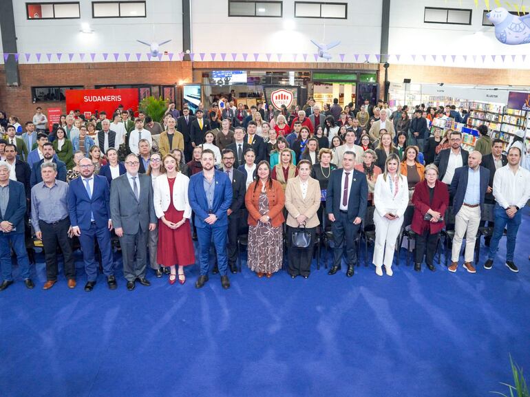 Gran grupo de aproximadamente 100 personas en trajes formales durante la inauguración en un ambiente interior decorado con estanterías de libros.