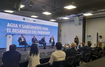 Cuatro panelistas, tres hombres en traje y una mujer con documento, discuten sobre saneamiento en un ambiente formal con público atento.