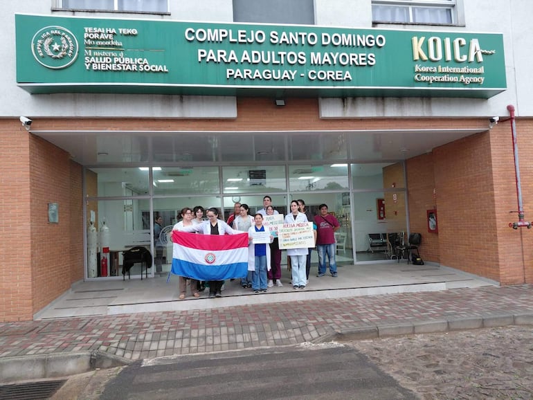 Seis personas en manifestación sosteniendo una bandera de Paraguay y carteles; mujer al frente lleva camiseta neutra y bandera.