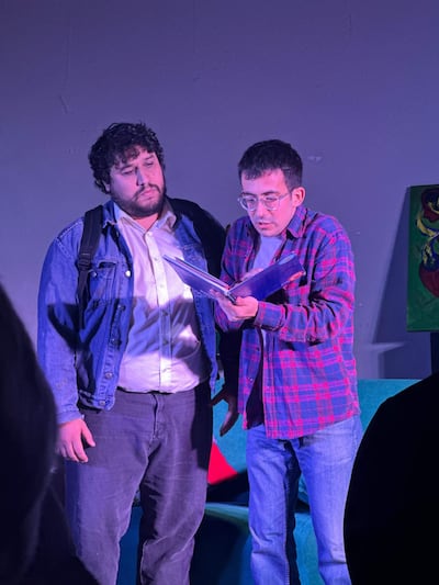Hombre con barba y chaqueta de mezclilla escucha a otro con gafas y camisa de cuadros, en un ambiente iluminado.